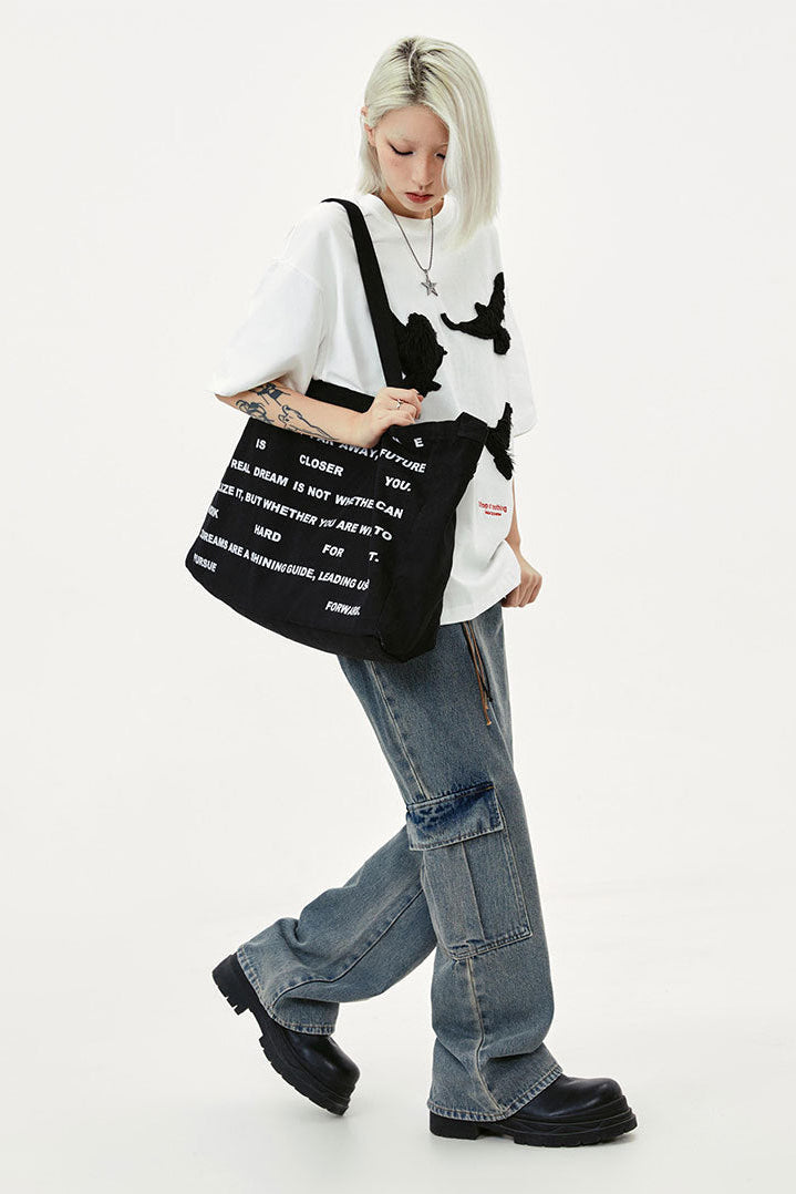 Side view of white dove embroidered T-shirt with a black text tote and wide-leg utility jeans