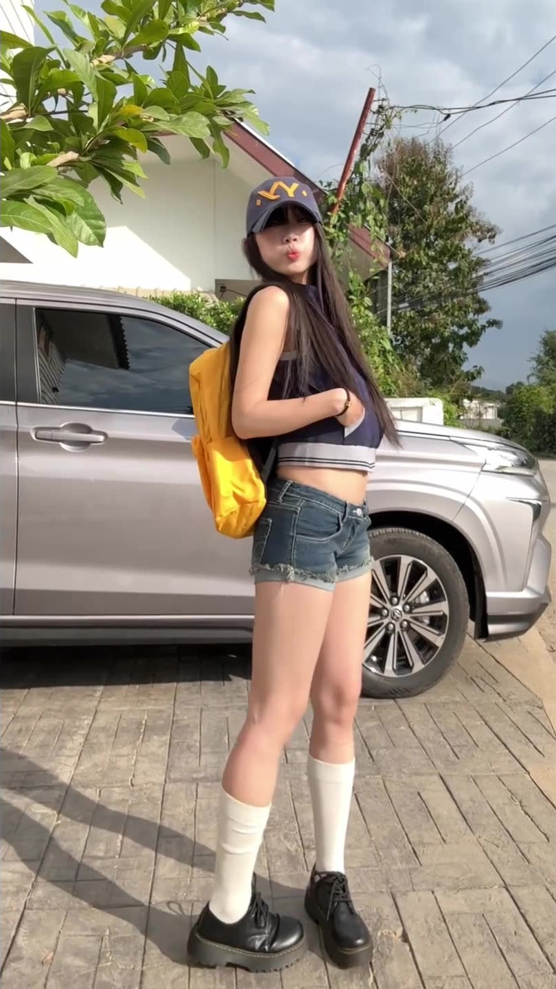 Model poses outdoors in raw hem denim shorts, crop top, and yellow backpack.