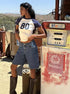 Woman in vintage gas station setting wearing dark wash knee-length denim shorts, white and navy ringer tee, and brown boots.