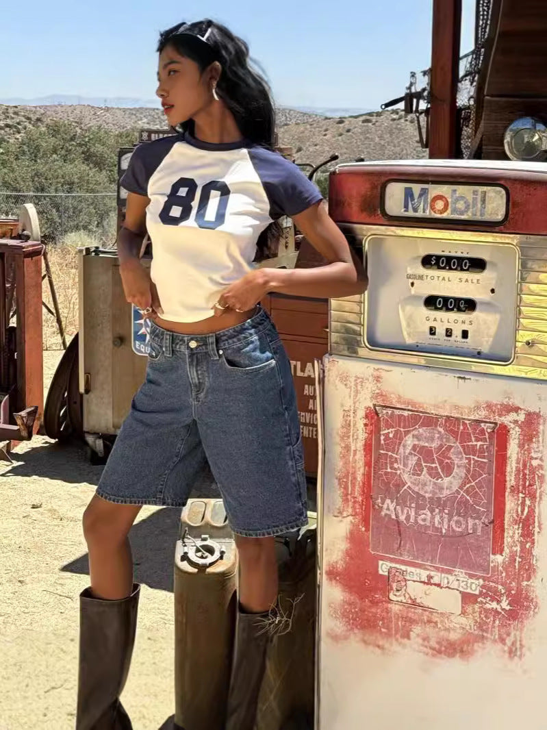 Woman in vintage gas station setting wearing dark wash knee-length denim shorts, white and navy ringer tee, and brown boots.
