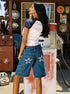 Woman showing back view of denim shorts with white star patches on pockets, standing by retro gas pump.