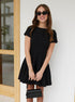 Smiling woman in black flared mini dress and sunglasses, holding a black purse.