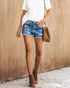 Full body shot of woman in distressed denim shorts, white t-shirt, brown heels, and a matching brown bag.