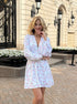 Woman smiling in white floral mini dress with v-neckline and long sleeves.