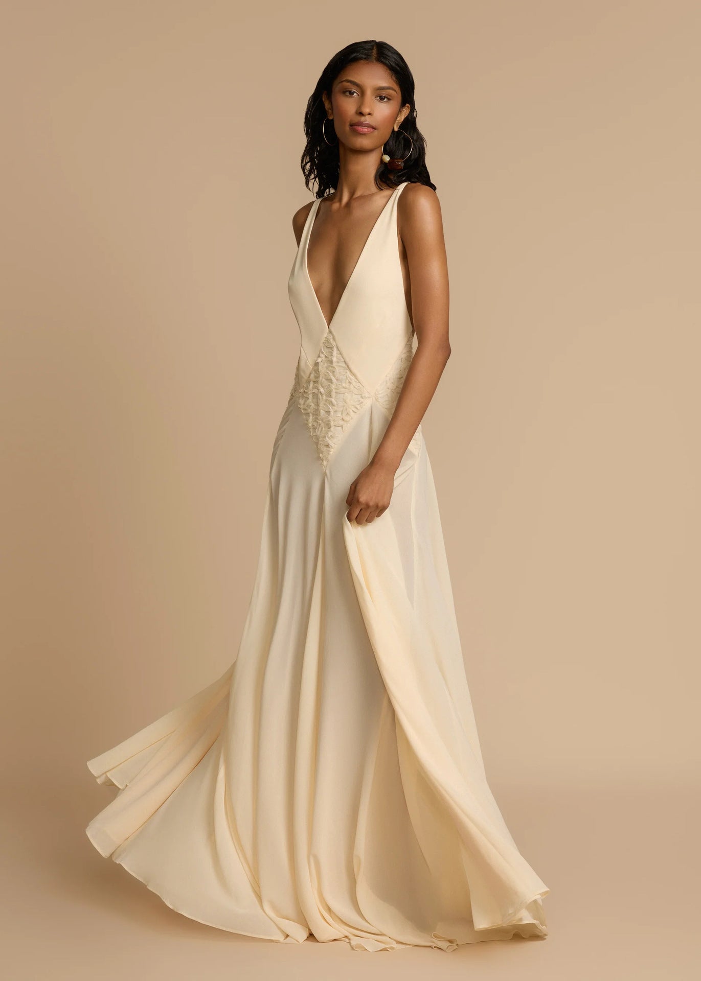 Woman wearing a white deep V vacation maxi dress with lace waist and flowing hem against a beige background