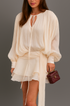 Close up of woman in a white chiffon ruffle set holding a small handbag against a gray background