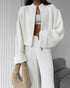 Person wearing a white lounge set with a cropped jacket and pants, holding a beige fur coat against a neutral background.