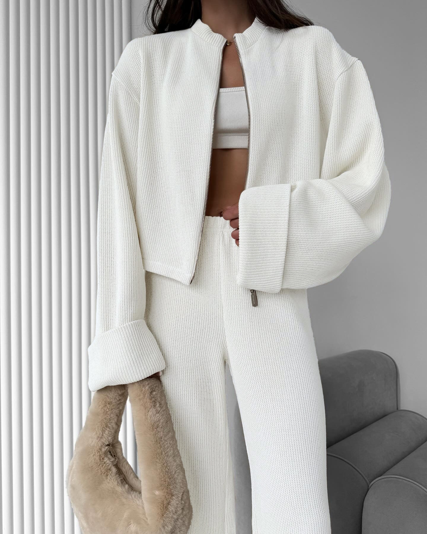 Person wearing a white lounge set with a cropped jacket and pants, holding a beige fur coat against a neutral background.