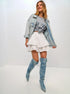 Woman smiling and posing in a light denim jacket, gray graphic tee, white ruffled mini skort, and blue tie-dye boots.