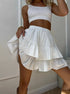 Woman wearing a white ribbed crop top and white ruffled polka dot mini skort, lifting one side of the skort to show the flounce detail.