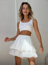 Woman smiling while wearing a white ribbed crop top and a white ruffled polka dot mini skort, standing against a white wall.