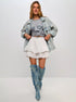 Woman posing in a light denim jacket, gray graphic tee, and white ruffled polka dot skort with blue tie-dye knee-high boots.