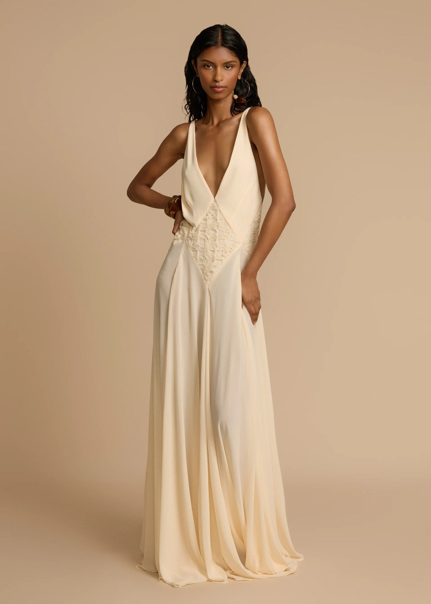 Woman wearing a white deep V vacation maxi dress with lace waist and flowing hem against a beige background