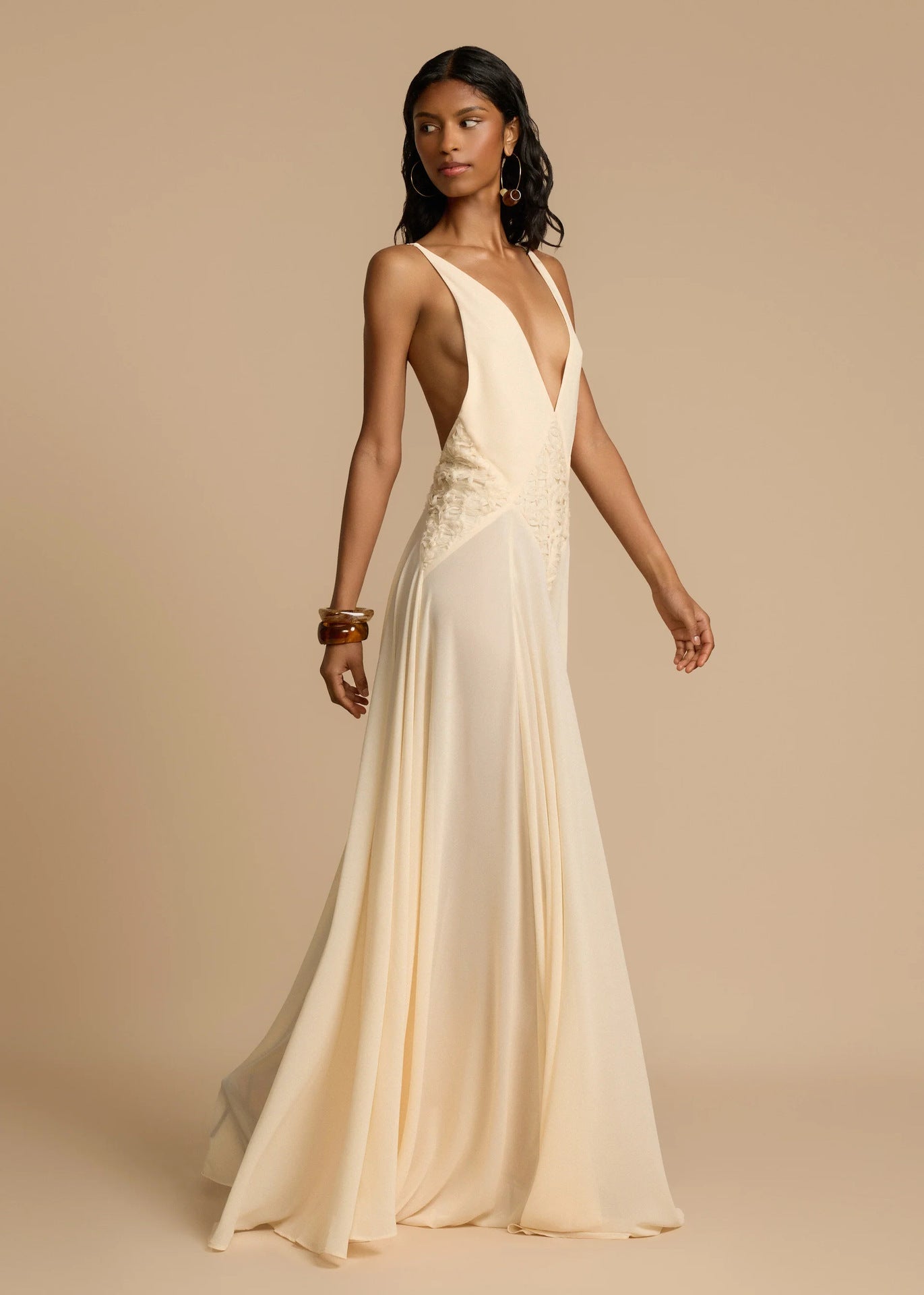 Woman wearing a white deep V vacation maxi dress with lace waist and flowing hem on a beige background