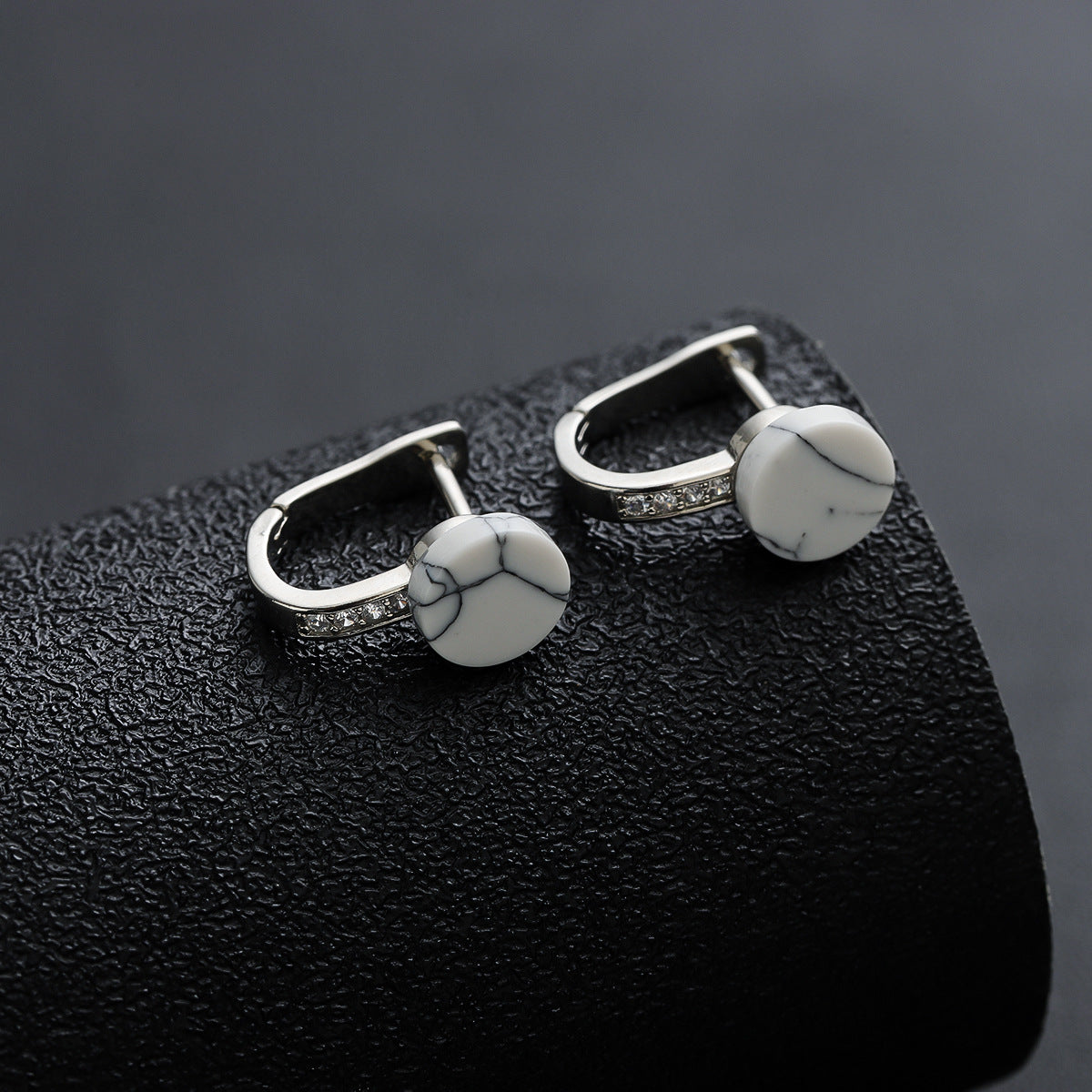 White marble-pattern stud earrings on textured black roll.