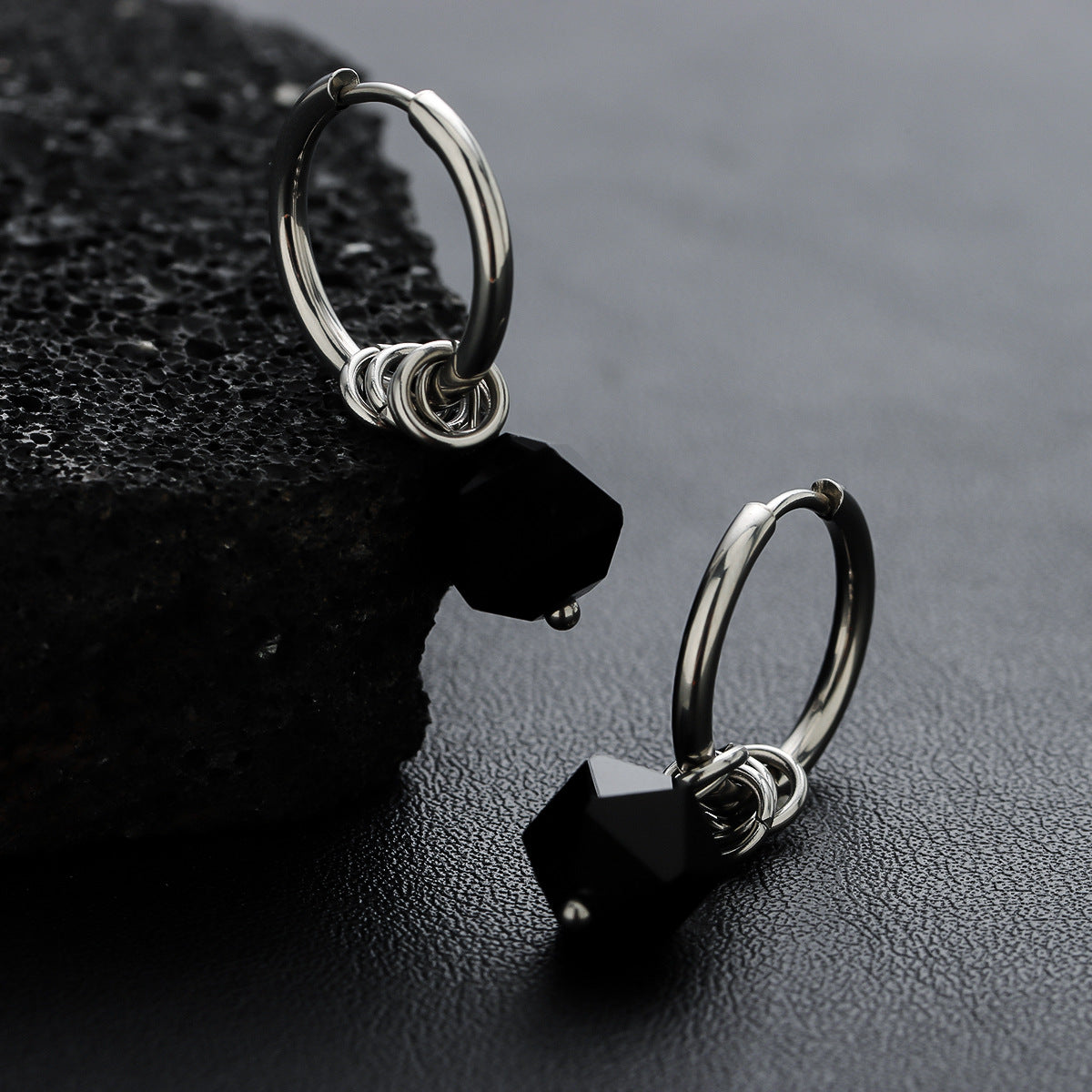 Silver hoop earrings with black hexagonal zircon drops beside stone.